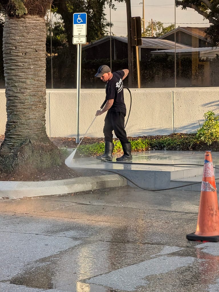 Technician pressure washing a commercial concrete walkway at sunset near a palm tree and handicap parking sign.
